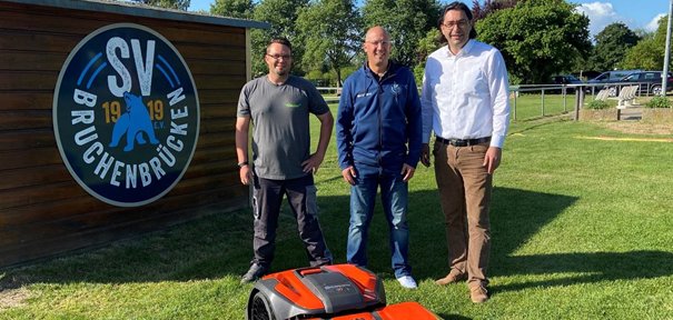 Foto: Bürgermeister Kjetil Dahlhaus (rechts) und Ralf Ringkowski vom SV Bruchenbrücken (mitte) nehmen den neuen Mähroboter für die Sportstätte Bruchenbrücken von der Firma Weimer Technik GmbH, Martin Dellmuth (links) in Empfang und starten die Inbetriebnahme.